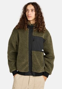 Veste en polaire vert olive avec un panneau avant en nylon noir, une poche zippée et des poignets côtelés. Texture douce avec une coupe décontractée.
