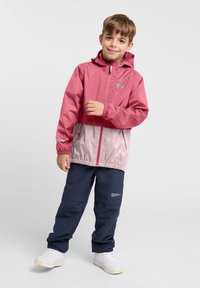 Rosa und graue Kapuzenjacke mit elastischen Bündchen, Reißverschluss und Logo. Kombiniert mit marineblauen Hosen und weißen Sneakern.