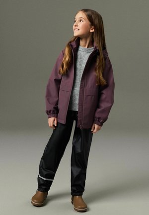 Mädchen mit langen Haaren, das eine lila Jacke mit Reißverschluss, einen grauen Pullover, schwarze wasserdichte Hosen mit reflektierenden Streifen und braune Stiefel trägt, schaut nach oben und lächelt.