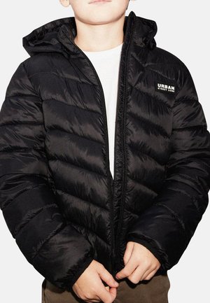 Enfant portant une doudoune noire à capuche avec le logo "Urban Street Zone", en train de fermer la fermeture éclair par-dessus une chemise blanche et un pantalon marron.