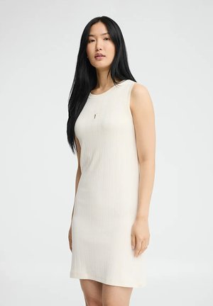 Giovane donna con lunghi capelli neri che indossa un abito crema senza maniche, lungo fino al ginocchio, con un piccolo logo ricamato sul petto.