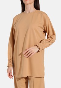 Lysebrune langærmede top fremstillet af glat stof med rund halsudskæring og samlede detaljer ved ærmekanterne.