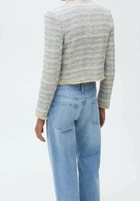 Veste grise texturée recadrée avec accents jaunes, associée à un jean denim bleu clair à coupe ample. Vue de dos mettant en évidence les bords effilochés.