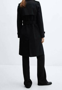 Trench-coat noir avec une taille cintrée par une ceinture, des poignets boutonnés, et un design arrière superposé, confectionné dans un tissu lisse. Associé à un pantalon noir.