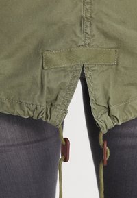 Olivgrüner Stoff mit gerafftem Saum, ausgestattet mit Kordelzügen und braunen Knebeln. Strukturierter, leichter Stoff für legere Kleidung.