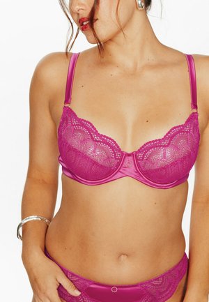 Femme portant un ensemble assorti de soutien-gorge et culotte en satin et dentelle magenta, avec des boucles d'oreilles et un bracelet argentés, posant devant un fond blanc.