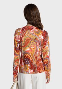 Blouse à manches longues avec un motif cachemire audacieux et coloré en rouge, orange et violet sur fond blanc, avec un col rond.