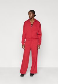 Červená mikina na zips a zodpovedajúce široké nohavice vyrobené z mäkkej tkaniny. Má predné vrecká a rebrované manžety. Čierne logo na hrudi.