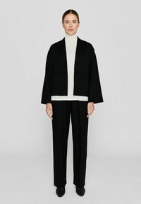 Zwarte oversized jas met brede mouwen, gedragen over een strakke witte coltrui. Gecombineerd met zwarte op maat gemaakte broeken en slanke zwarte schoenen.