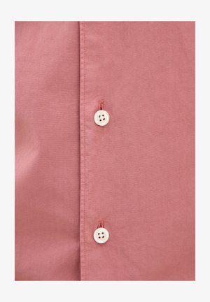 Vaaleanpunainen puuvillapaita, jossa on sileä pinta. Paidassa on kaksi valkoista nappia, joissa on neljä reikää kummassakin, tasaisesti napinläpiväliin sijoitettuna.