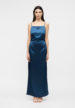 Moteris ilgais tamsiais plaukais, dėvinti mėlyną satino maxi suknelę be rankovių su plonomis petnešėlėmis ir baltus atvirus aukštakulnius batelius, stovinti priešais šviesų vienodo fono.