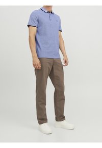 Man med ljusblå pikétröja, bruna byxor och vita sneakers som står mot en enkel ljus bakgrund, huvudet är inte synligt.