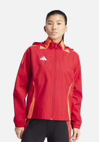 Veste imperméable rouge avec des accents orange, fermeture éclair intégrale à l'avant, capuche et poignets élastiques. Présente un logo sur le côté supérieur gauche. Design léger.