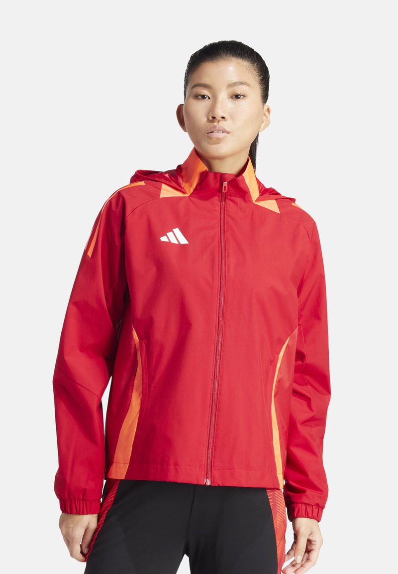 Veste imperméable rouge avec des accents orange, fermeture éclair intégrale à l'avant, capuche et poignets élastiques. Présente un logo sur le côté supérieur gauche. Design léger.