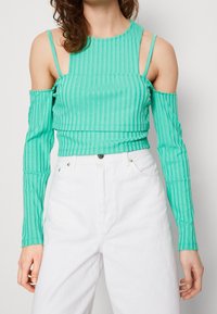 Nainen, jolla on päällään teal-vihreä ribbipinta kyljistä avattu yläosa, pitkät hihat ja valkoiset vyötäröä korostavat housut, seisoo vihreää taustaa vasten.