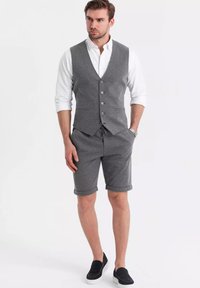 Gilet grigio a scacchi con cinque bottoni sopra una camicia bianca a maniche lunghe, abbinato a pantaloni corti grigi. Scarpe nera senza lacci completano il look.