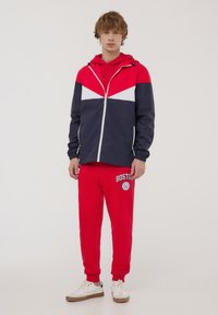 Giacca a vento rossa e blu navy con accenti bianchi, chiusura con zip e cappuccio. Abbinata a pantaloni sportivi rossi con grafica "BOSTON" e sneakers bianche.