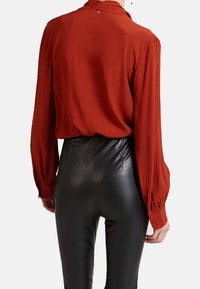 Hnědý top v odstínu rzi s dlouhými rukávy a knoflíkovými manžetami, doplněný černými kalhotami z umělé kůže s vysokým pasem a hladkou texturou.