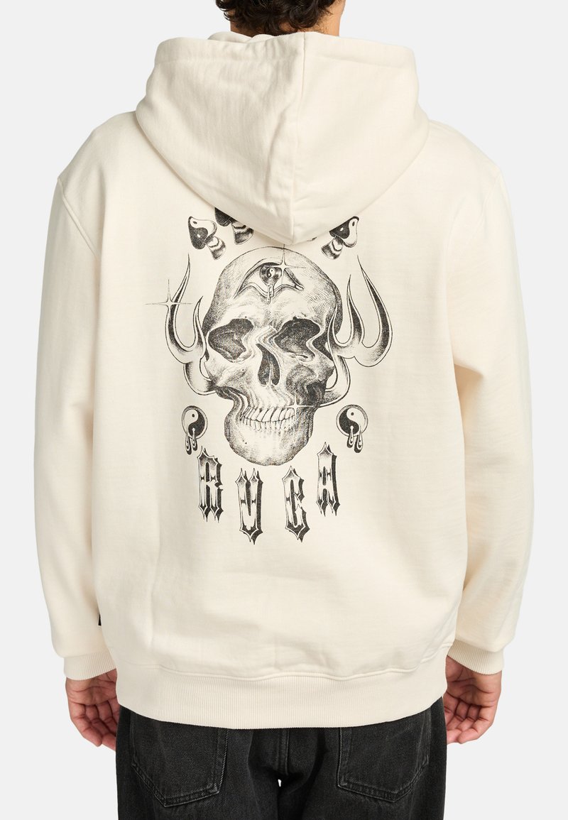 Persoon in een crèmekleurige hoodie met een zwart schedelontwerp, inclusief een derde oog, drietanden, yin-yang symbolen en tekst in gotische stijl op de achterkant.