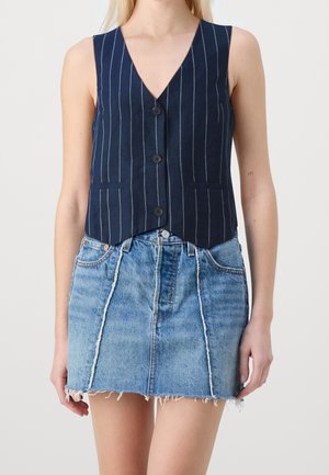 Kvinde iført en ærmeløs navyblå vest med knapper og søm i striber, kombineret med en lyseblå denim mininederdel med frynser.