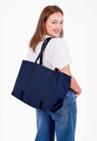 Tyrkysová taška přes rameno vyrobená z odolné tkaniny, s dvěma širokými popruhy a jemným logem, má prostorný obdélníkový tvar.