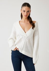 Hvid strikket sweater med en dyb V-udskæring, der har struktureret rib og kabelmønstre. ærmerne er brede og let oversized.