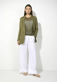 Olivgrüner Blazer über einem Netzstricktop, kombiniert mit lockeren weißen Leinenhosen. Merkmale sind der lässige Schnitt und der leichte Stoff, dazu offene Sandalen.