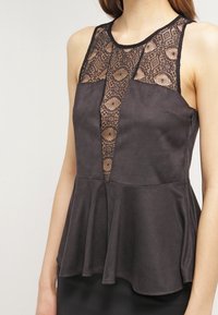 Top peplum negro con inserto de encaje de patrón geométrico, cuello alto, diseño sin mangas y textura de tela suave.