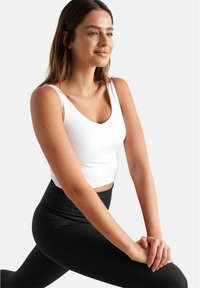 Valge sobiv tank top, millel on sügava kaelusega lõige, koos kõrge talje mustade retuusidega. Kanga tekstuur näib olema sile ja veniv.
