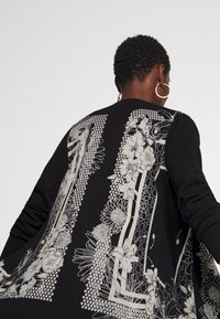 Svart kofta med detaljerat vitt blommönster och geometriskt mönster, med avslappnad passform och ribbade muddar. Mjuk textur, lättviktsmaterial.