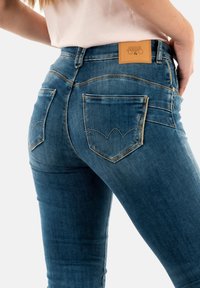 Donkerblauwe denimjeans met een soepele textuur. Opmerkelijke kenmerken zijn een leren label op de tailleband en afwijkende stiksel op de achterzak.