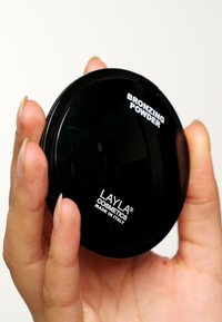 Svart rund kompakt med bronzerpulver, märkt "BRONZING POWDER" och "LAYLA COSMETICS MADE IN ITALY," hålls i en hand.