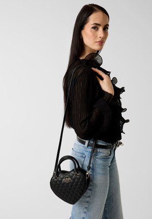 Frau trägt eine schwarze, durchsichtige Bluse mit Rüschen und hellblaue Jeans, sie trägt eine schwarze, gesteppte, herzförmige Guess-Handtasche mit Schulterriemen.