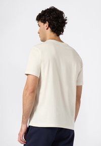 Jonge man met gekruld donker haar, gekleed in een eenvoudig off-white T-shirt met korte mouwen en donkere broek, met zijn rug naar de camera gekeerd tegen een witte achtergrond.