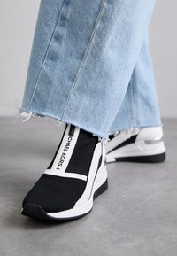 Sneaker in tessuto nero con accenti bianchi, caratterizzato da un logo prominente, pannelli laterali elastici e una suola testurizzata. Abbinato a jeans azzurri sfilacciati.