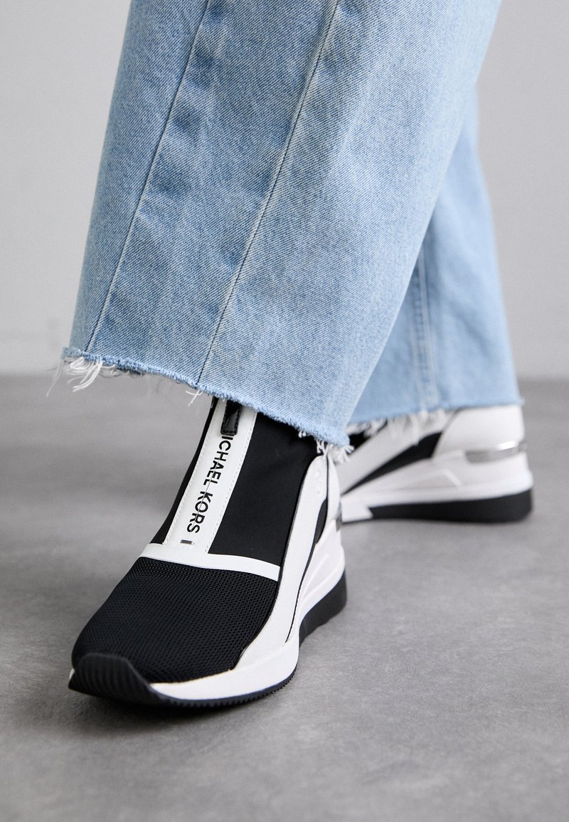 Zapatillas de tela negras con acentos blancos, que presentan un logotipo prominente, paneles laterales elásticos y una suela texturizada. Combinadas con jeans de mezclilla claros y deshilachados.