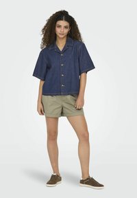 Camicia di jeans a maniche corte, con chiusura frontale a bottoni, dettagli di cuciture chiare, abbinata a pantaloni corti khaki e sneaker marroni.