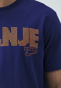 T-shirt en coton bleu marine avec motif de points orange et texte "ORANJE", accompagné d'un logo Nike et de coordonnées imprimées en dessous. Modèle à manches courtes.