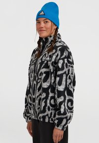 Vrouw met gevlochten haar, gekleed in een blauwe beanie en een zwart-wit gestreepte fleecejack, staand tegen een effen achtergrond.