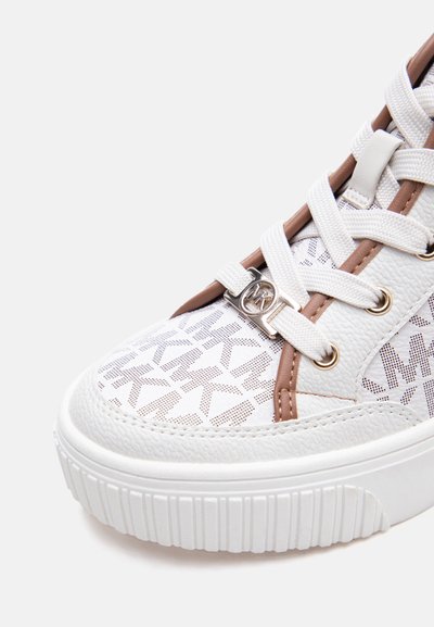 Λευκό μπροστινό μέρος αθλητικού παπουτσιού με υφασμένο πέλμα, λευκά κορδόνια, καφέ περίγραμμα και διακοσμητικό πάνω μέρος με επαναλαμβανόμενα γράμματα "MK" και ασημένιο μενταγιόν κορδονιών MK.