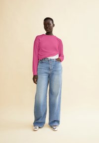 Rosa Langarmshirt mit lässigem Schnitt, kombiniert mit hellblauen weit geschnittenen Jeans. Das Modell steht vor einem neutralen Hintergrund und trägt silberne Schuhe.