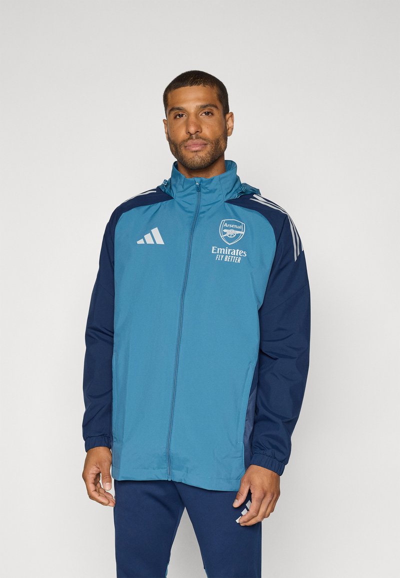 Blauwe en marineblauwe jas met ritsluiting, capuchon en elastische manchetten; voorzien van Adidas- en Arsenal-logo's, lichtgewicht materiaal en zijzakken.