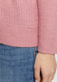 Pull en tricot rose avec un motif texturé et une étiquette portant "QS". Associé à un denim bleu, mettant en valeur une coupe décontractée.