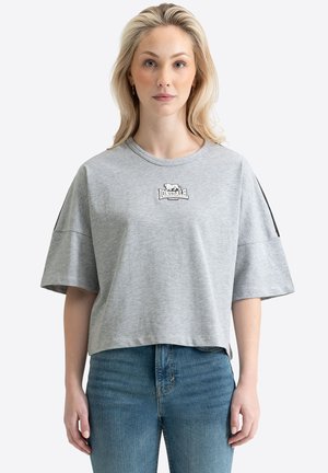 Mujer rubia con camiseta cortada de Lonsdale London gris clara con rayas negras en las mangas, combinada con vaqueros azules, mirando hacia adelante.