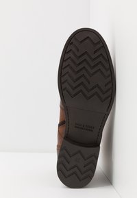 Bottine en cuir marron avec une semelle en caoutchouc présentant un motif de bande de roulement en zigzag. Marquage de la marque et taille "42" estampillés sur la semelle.