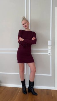 Kvinne med blondt hår i en knute, iført en mørk lilla kjole og svarte ankelstøvler, stående med armene i kors foran en grå panelvegg.