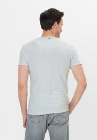 Uomo visto di spalle che indossa una camicia azzurro chiaro a maniche corte e jeans grigi, in piedi contro uno sfondo bianco uniforme.