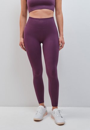 Femme portant des leggings violets taille haute et un soutien-gorge de sport assorti avec des baskets blanches sur un fond clair uni.