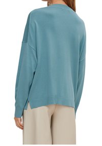 Femme portant un pull ample teal avec des poignets et un ourlet côtelés, associé à un pantalon beige clair à jambes larges, vue de dos.
