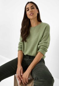 Maglione verde chiaro, morbido, con texture, collo rotondo e maniche arrotolate, abbinato a pantaloni in velluto a coste verde scuro. Design minimalista.